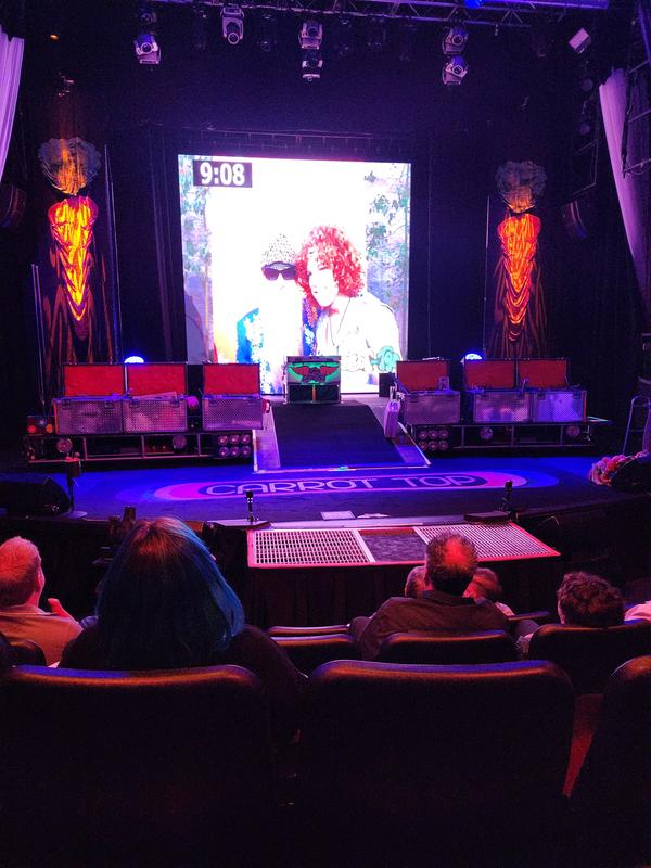 a group of people sitting in chairs in front of a stage with a large screen - Where to sit when seeing Carrot Top?